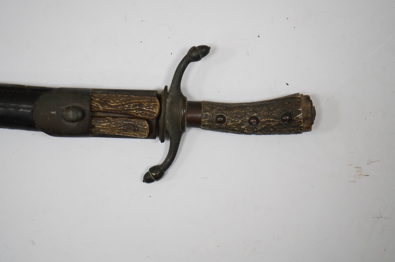 A 19th century Continental hunting hanger, blade cut with twin fullers, recurved brass crosspiece with acorn finials, staghorn grip (pommel missing), in its brass mounted leather scabbard, containing knife and fork (chap